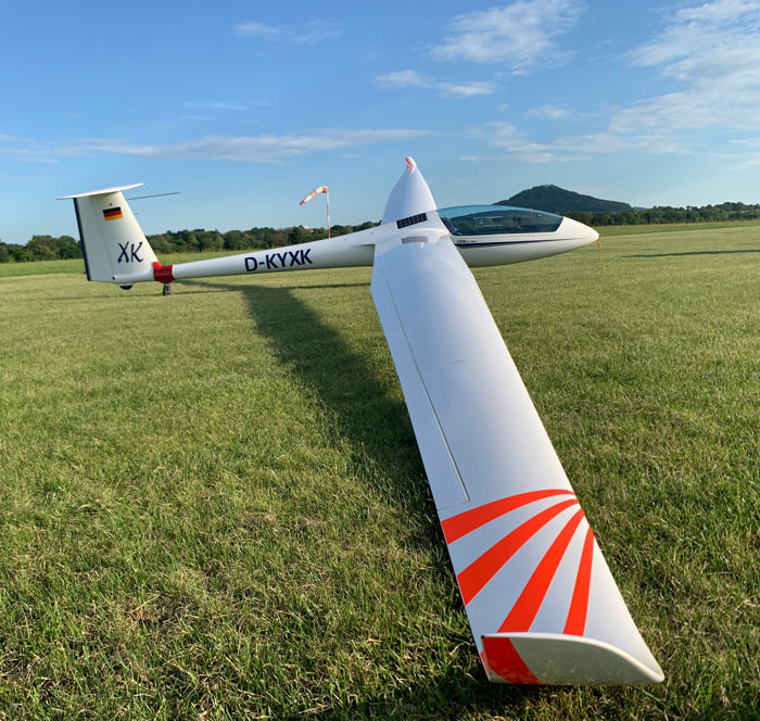 White glider sailplane with long wing on grassy airfield, windsock and hill under blue sky.
